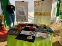 Para cego ver: cenário composto por mesa com produtos feitos pelo projeto. AO fundo, um banner do Mulheres Mil e outro do Produzir para Libertar e um manequim vestido com camiseta produzida pelo projeto