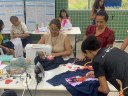 Para cego ver: mulheres distribuídas em uma sala, costuram roupas. Ao centro, uma com máquina de costura e ao redor delas, mulheres costuram a mão